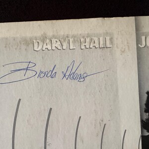 May include: A black and white photo of a person with the text "JOHN" above their head. The photo is signed "Brenda Adams" and there is text that says "Daryl Hall" above the signature.