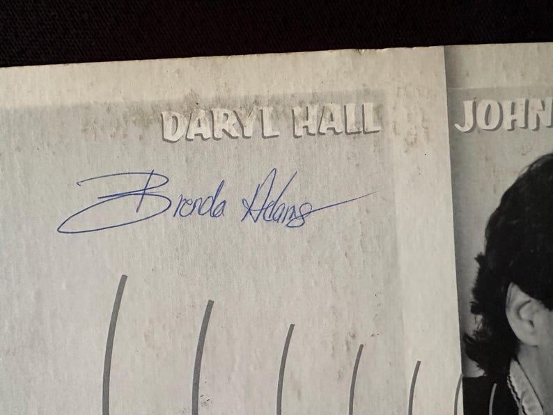 May include: A black and white photo of a person with the text "JOHN" above their head. The photo is signed "Brenda Adams" and there is text that says "Daryl Hall" above the signature.
