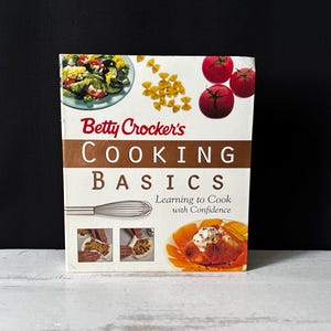 May include: A cookbook titled "Betty Crocker's Cooking Basics" with images of food, including tomatoes, pasta, and a finished dish. The cover features the title in large, bold letters and a whisk illustration.