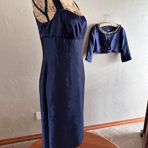 May include: A navy blue formal dress with a matching bolero jacket. The dress features a square neckline and a fitted silhouette. The bolero has a beaded collar. The dress is displayed on a mannequin.