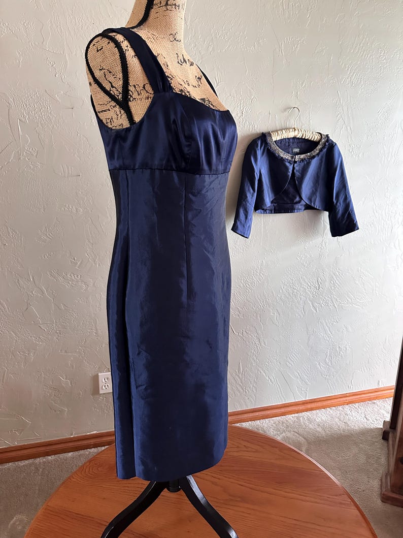 May include: A navy blue formal dress with a matching bolero jacket. The dress features a square neckline and a fitted silhouette. The bolero has a beaded collar. The dress is displayed on a mannequin.