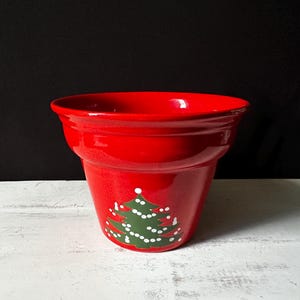 May include: A red ceramic planter featuring a green Christmas tree design. The tree has white dots, resembling ornaments.
