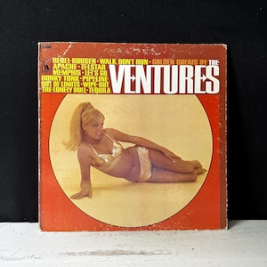 May include: A red and yellow vinyl record album cover featuring a woman in a gold bikini lying on a beach. The album title is "Golden Greats by The Ventures" and lists songs like "Rebel Rouser", "Walk Don't Run", "Apache", "Telstar", "Memphis", "Let's Go", "Honky Tonk", "Pipeline", "Out of Limits", "Wipe Out", "The Lonely Bull", and "Tequila".