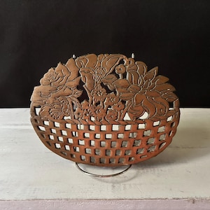 May include: A brown, oval decorative basket with a floral design carved into the top. The basket has a lattice-style body and is supported by a metal stand. The floral design includes roses and other flowers.