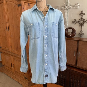 Puede incluir: Una camisa de mezclilla azul claro de manga larga con cierre de botones y dos bolsillos en el pecho. La camisa tiene un aspecto descolorido y una pequeña etiqueta en la parte inferior izquierda que dice "Tommy Hilfiger".