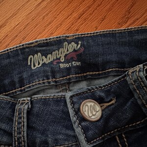 Pode incluir: Close-up de jeans Wrangler Boot Cut azul escuro. Os jeans t&ecirc;m um fecho de bot&atilde;o com o logotipo Wrangler. A palavra "Wrangler" &eacute; bordada em verde claro com um acento rosa acima da palavra "Boot Cut".