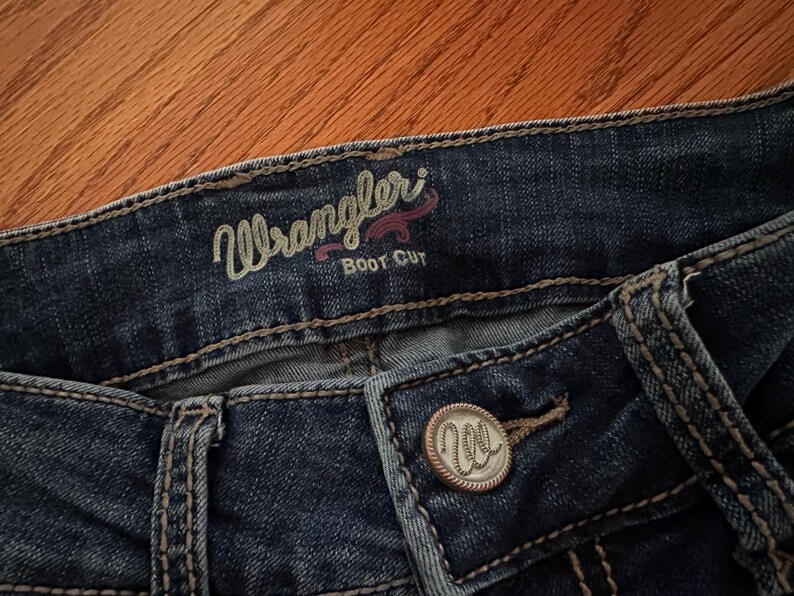 Pode incluir: Close-up de jeans Wrangler Boot Cut azul escuro. Os jeans t&ecirc;m um fecho de bot&atilde;o com o logotipo Wrangler. A palavra "Wrangler" &eacute; bordada em verde claro com um acento rosa acima da palavra "Boot Cut".
