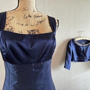 May include: A navy blue formal dress with a square neckline and wide straps is displayed on a mannequin. A matching bolero jacket with a beaded collar hangs nearby. The dress and jacket are made of a shiny fabric.