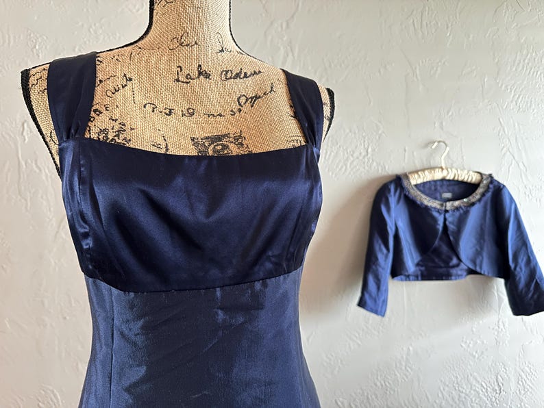 May include: A navy blue formal dress with a square neckline and wide straps is displayed on a mannequin. A matching bolero jacket with a beaded collar hangs nearby. The dress and jacket are made of a shiny fabric.