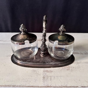 May include: A silver-toned condiment server featuring two clear glass bowls with matching silver-toned lids. The set is connected by a central vertical handle attached to an oval base, with small notches in the lids for spoons to rest inside.