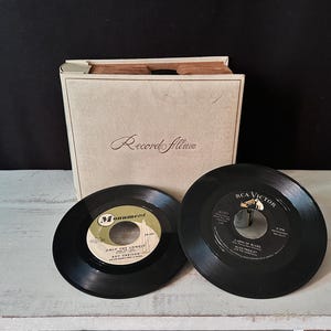 May include: A vintage off-white record album storage case with the text Record Album printed on the front. Two black 45 RPM vinyl records are positioned in front of the case, one featuring a Monument label and one with an RCA Victor label.