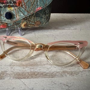 Può includere: Occhiali vintage a occhi di gatto con montatura rosa e trasparente. Gli occhiali hanno uno stile retrò, con un'asta marrone chiaro e un astuccio abbinato sullo sfondo. La custodia ha un motivo floreale e il testo "OKLA. CITY, OKLA."