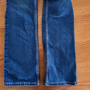 Pode incluir: Close-up da parte inferior de um par de jeans de ganga azul. Os jeans t&ecirc;m um design cl&aacute;ssico de cinco bolsos com costuras vis&iacute;veis ao longo das costuras e bainhas. O denim tem uma lavagem m&eacute;dia com desbotamento sutil.