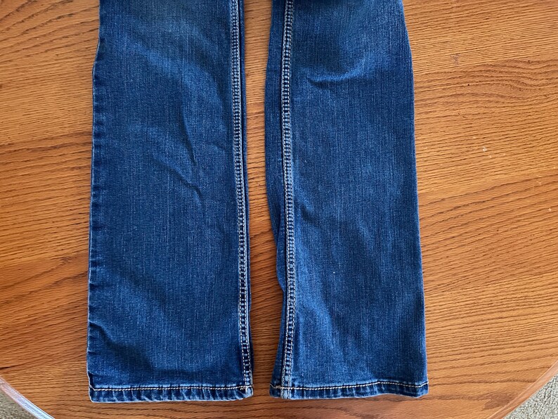 Pode incluir: Close-up da parte inferior de um par de jeans de ganga azul. Os jeans t&ecirc;m um design cl&aacute;ssico de cinco bolsos com costuras vis&iacute;veis ao longo das costuras e bainhas. O denim tem uma lavagem m&eacute;dia com desbotamento sutil.