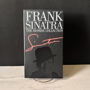 Puede incluir: Una portada de libro negra con el título "FRANK SINATRA" en letras blancas. Debajo, en letras blancas más pequeñas, se lee "THE REPRISE COLLECTION". El nombre "Sinatra" está escrito en rosa. Una silueta de Frank Sinatra está en la parte inferior.