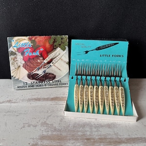 Puede incluir: Un juego de 12 tenedores de acero inoxidable para aperitivos en una caja etiquetada como Little Fork. Los tenedores tienen mangos de color blanco roto con un patrón floral. El empaque muestra un plato de comida y el texto Special Quality Little Forks.