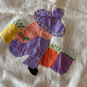 May include: A purple, orange, and yellow patchwork appliqué of a person wearing a hat and carrying two bags. The appliqué is stitched onto a white fabric background.