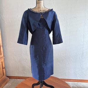 May include: A navy blue dress and matching jacket set displayed on a mannequin. The dress is a sheath style, and the jacket has three-quarter sleeves and a fringed neckline. The outfit is made of a smooth, possibly silk-like fabric.