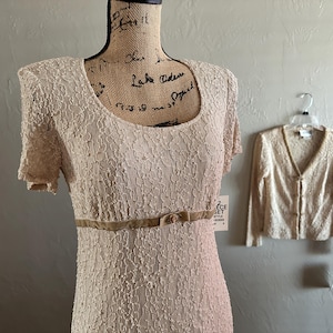 Può includere: Un abito in pizzo beige con scollo tondo e maniche corte è esposto su un manichino. Un cardigan in pizzo abbinato è appeso a un gancio a destra. L'abito presenta una cintura in velluto marrone. Il set è un completo di due pezzi.