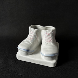 May include: A white ceramic decorative piece shaped like ice skates. The skates have pink laces and blue accents on the bottom. The piece is on a white rectangular base.