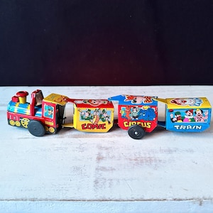 Könnte beinhalten: Ein farbenfroher Vintage-Spielzeugzug mit roten, blauen und gelben Wagen. Die Wagen sind mit "COMIC", "CIRCUS" und "TRAIN" beschriftet und zeigen Cartoon-Illustrationen. Das Spielzeug hat schwarze Räder und steht auf einer weißen Oberfläche.
