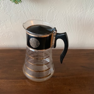 May include: A black and gold glass coffee pot with a black lid and a black handle. The pot has a gold band around the middle and a gold design on the lid.