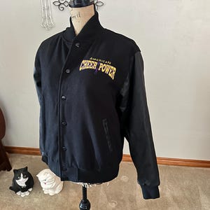 以下が含まれることがあります： 黒いスタジアムジャンパー。袖は黒で、「AMERICAN CHEER POWER」の文字が金と紫で書かれています。ボタン留めのフロントとリブ編みの襟と袖口が付いています。クラシックなスポーツウェアです。