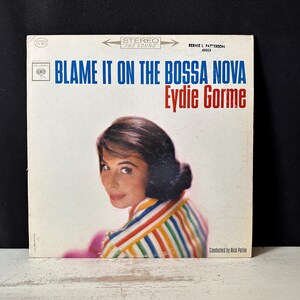Könnte beinhalten: Ein Vintage-Vinyl-Schallplattencover mit dem Titel Blame It On The Bossa Nova von Eydie Gorme. Das Cover zeigt ein Porträt einer Person mit dunklem Haar in einem mehrfarbig gestreiften Hemd, mit blauem und rotem Text auf weißem Hintergrund.