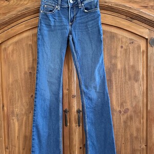 Pode incluir: Jeans Wrangler azuis com design de perna flare. Os jeans est&atilde;o pendurados em um cabide preto contra um fundo de madeira. Os jeans t&ecirc;m fecho de bot&atilde;o e z&iacute;per, com bolsos e costuras decorativas.
