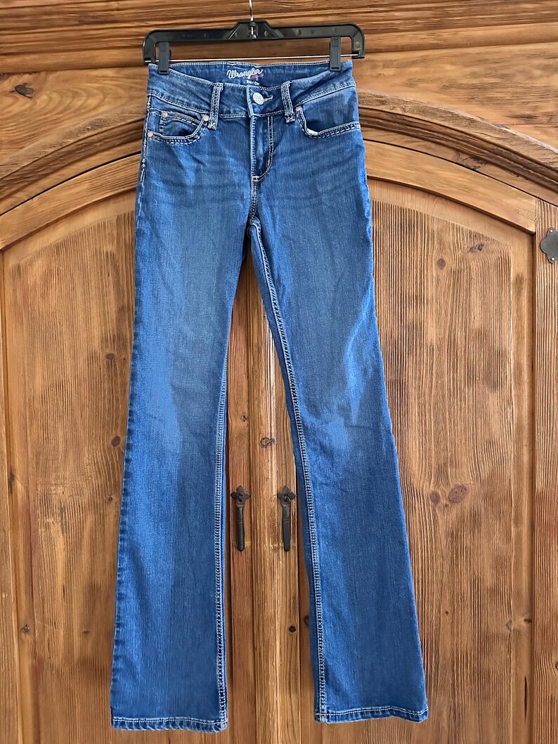 Pode incluir: Jeans Wrangler azuis com design de perna flare. Os jeans est&atilde;o pendurados em um cabide preto contra um fundo de madeira. Os jeans t&ecirc;m fecho de bot&atilde;o e z&iacute;per, com bolsos e costuras decorativas.