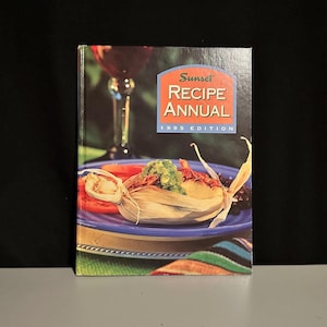 May include: The cover of the "Sunset Recipe Annual 1995 Edition" cookbook showcases a colourful food arrangement. A tamale, tomatoes, and black beans are arranged on a blue plate. A wine glass is visible in the background, suggesting a culinary theme.