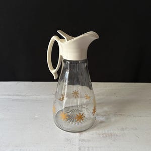 May include: A clear glass jug with a white plastic lid and a pour spout. The jug has a gold star pattern and is designed for serving syrup or sauce.