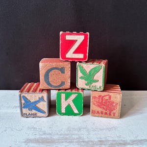 May include: A stack of vintage wooden alphabet blocks. The blocks are painted with letters, images, and words in various colors. The letters include Z, C, K. Images include a plane, eagle, and a truck.