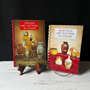 Puede incluir: Dos libros encuadernados en espiral con ilustraciones coloridas de cerámica. El libro de la izquierda se titula "Weller Art Pottery in Color" y el libro de la derecha se titula "Roseville Art Pottery in Color".