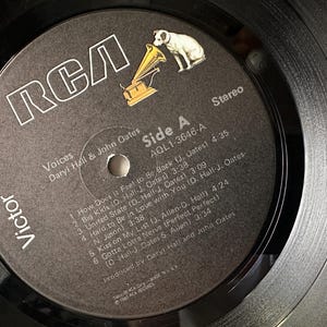 May include: A black and white vinyl record with a black label. The label has the RCA logo, the title "Voices" by Daryl Hall & John Oates, and the track list for side A of the record. The record is in stereo and has the catalog number AQL1-3646-A.