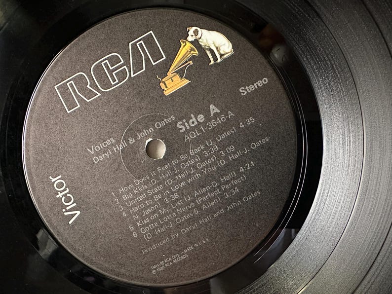 May include: A black and white vinyl record with a black label. The label has the RCA logo, the title "Voices" by Daryl Hall & John Oates, and the track list for side A of the record. The record is in stereo and has the catalog number AQL1-3646-A.