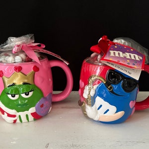Puede incluir: Dos tazas de personajes de M&M's con temática de San Valentín. La taza rosa presenta un M&M verde vestido de reina con una corona dorada y el texto "My Queen". La taza roja presenta un M&M azul con gafas de sol y tocando el saxofón. Ambas tazas tienen una tapa de plástico transparente y están llenas de caramelos M&M's.