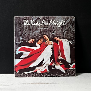 Könnte beinhalten: Ein Vintage-Vinyl-Plattencover von The Who. Das Albumcover ist schwarz mit weißem Text, der "The Kids Are Alright" und "The Who" lautet. Das Albumcover zeigt ein Foto von drei Männern, die auf einer rot-weiß-blauen Decke liegen.
