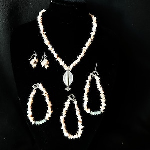Puede incluir: Un conjunto de joyas que incluye un collar, pendientes y dos pulseras, todos hechos con pequeñas astillas de piedra rosa claro y blanca. El collar tiene un colgante plateado. Las joyas se exhiben sobre un fondo de terciopelo negro.