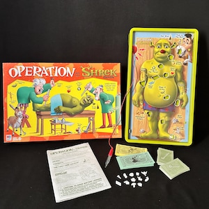 May include: A Shrek-themed Operation board game. The game features a green Shrek figure with various body parts to remove, a game board, cards, and play money. The box art shows Shrek on an operating table.