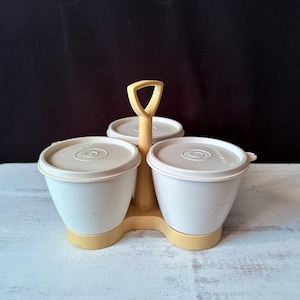 Puede incluir: Un juego de condimentos de cocina vintage con tres recipientes blancos con tapas, colocados en un soporte amarillo pálido con asa. Los recipientes tienen forma redondeada y probablemente son de plástico.
