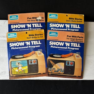 May include: Four vintage "Show 'N Tell" record players with different themes: "Fun With Clocks", "Bible Stories Noah Builds An Ark", "Bible Stories David Meets Goliath", and "Fun With Facts The Wright Brothers' Flying Machine". Each player features a colorful picture on the front and a record player with a spinning record.
