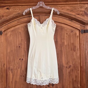 May include: A pale yellow slip dress featuring a V-neckline with white floral lace trim, thin adjustable shoulder straps, and a matching white lace hemline. The garment has a fitted bodice and a straight skirt silhouette with a smooth finish.