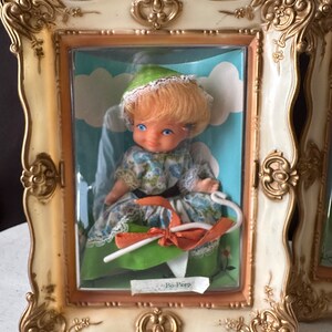 May include: A vintage Bo-Peep doll in a white dress with a green and blue floral pattern, wearing a green hat and holding a white shepherd's crook. The doll is displayed in a gold and white ornate frame with a clear plastic cover.