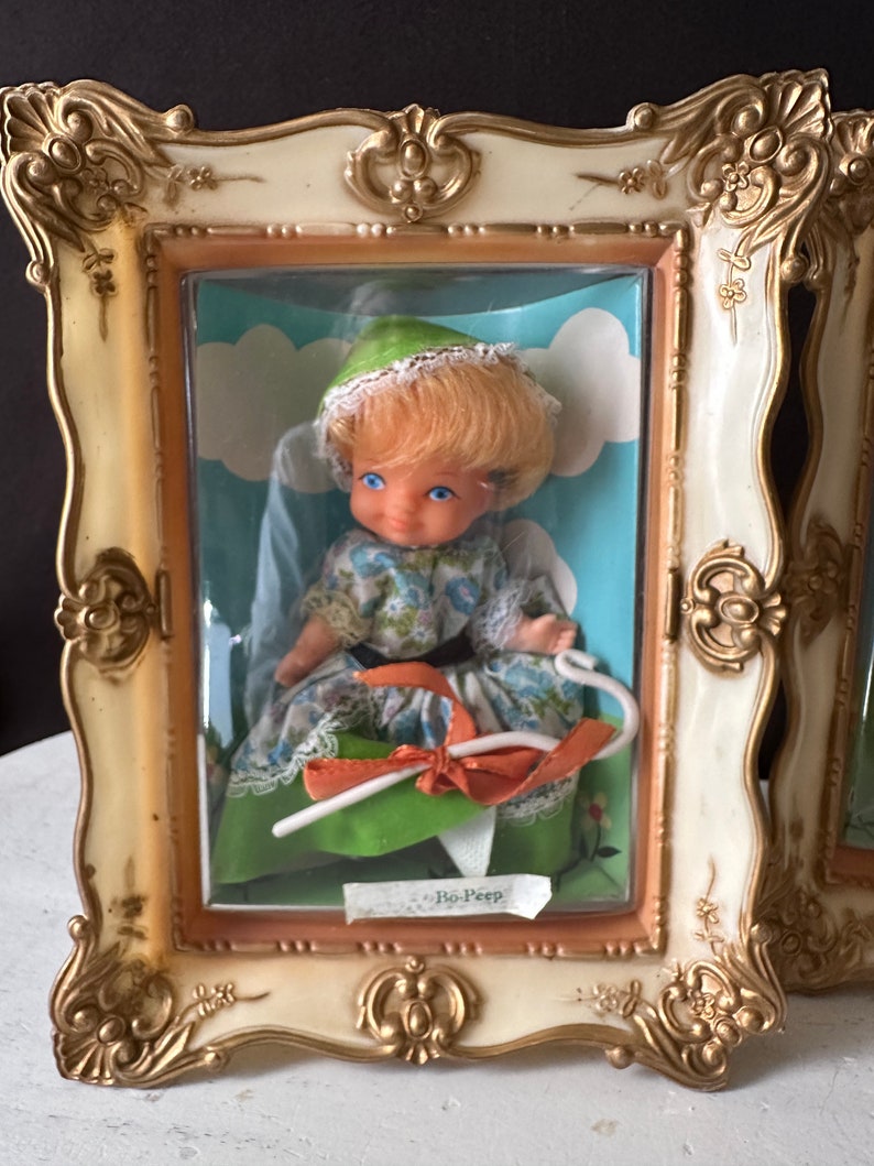May include: A vintage Bo-Peep doll in a white dress with a green and blue floral pattern, wearing a green hat and holding a white shepherd's crook. The doll is displayed in a gold and white ornate frame with a clear plastic cover.