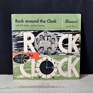 May include: A vintage vinyl record sleeve for Rock around the Clock by Bill Haley and his Comets on the Brunswick label. The cover features a light green background, a black and white photo of the band, and a clock graphic with the text ROCK CLOCK.