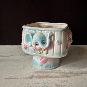 May include: A vintage ceramic planter with a light blue and white color scheme. The planter features an elephant and a girl's face on opposite sides. The elephant has large ears and a yellow bow tie. The planter is on a pedestal base.