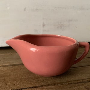 Puede incluir: Una salsera de cerámica rosa con pico vertedor y asa. La salsera es de color liso y se encuentra sobre una superficie de madera. El fondo es una pared de madera pintada de blanco.