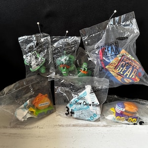 Puede incluir: Una colección de juguetes de plástico en bolsas de plástico transparentes. Los juguetes incluyen un monstruo verde, un superhéroe azul y rojo, un tren amarillo y verde y un tren azul y amarillo. Las bolsas tienen texto en ellas, incluyendo "Wacky Chew Chew Train", "Sonic", "Sports Gear" y "Incredible Munchers".
