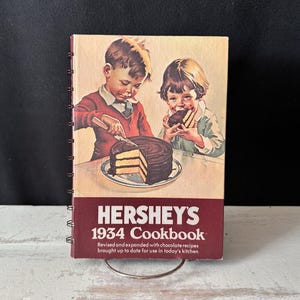 Op de afbeelding: Een spiraalgebonden Hershey's 1934 Cookbook met een illustratie van twee kinderen die een chocoladetaart eten. De kaft bevat de tekst: HERSHEY'S 1934 Cookbook, Revised and expanded with chocolate recipes brought up to date for use in today's kitchen.